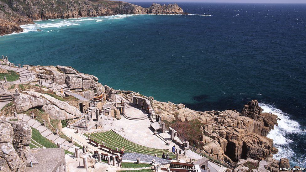 This image shows the current-day Minack Theatre. There is a concrete walkway leading down the cliff which has rope handrails. The walkways are surrounded by grass and the seats are also grass. There is a stage made of stone with a stone window scene facing the sea. To the left of the stage are balcony seats with stone pillars in front and the pillars are visible on either side of the stage. The has granite boulders to the left, as well as beneath the venue along the waters edge. In the background is the sea