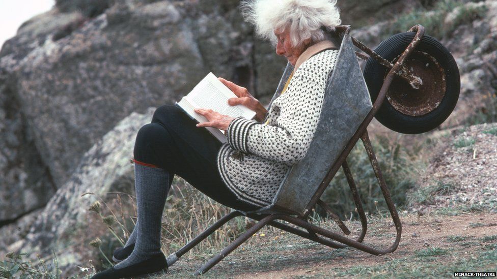 This images shows an older Rowena Cade leaning back in a silver wheelbarrow whilst reading a thick book. Her white hair looks wild as the wind blows through it and she is wearing a white woollen cardigan, covered in a pattern of black squares along with a black and white striped trim along the sleeves and waste. She is wearing black jodhpurs and knee length grey woollen socks with an orange trim, as well as black plimsols. In the background, there are granite boulders and Cade is on a patch of grass.