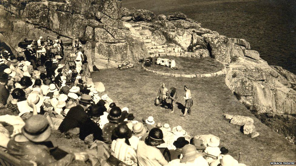 This image displays the very first performance at the Minack Theatre in 1932. The image is in sepia tones and shows three performers in costumes on the grassy stage, with the granite boulders of the Minack Headland in the background. The audience is wearing an array of sunhats, formal dresses and suits as they watch from the seats overlooking the makeshift stage. In the background is the sea looking dark against the lit stage.