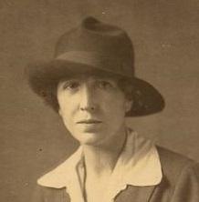 This images shows Katharine Burdekin. She is wearing a blazer with a white collared shirt in a V-neck, accompanied with a fedora tilted diagonally from the left. The image is in sepia tones. Burdekin has a slender, oval face dark eyes and short, dark curly hair tucket into her hat. 